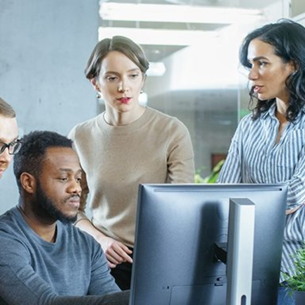 A man and women surrounding a man sitting next to the computer
