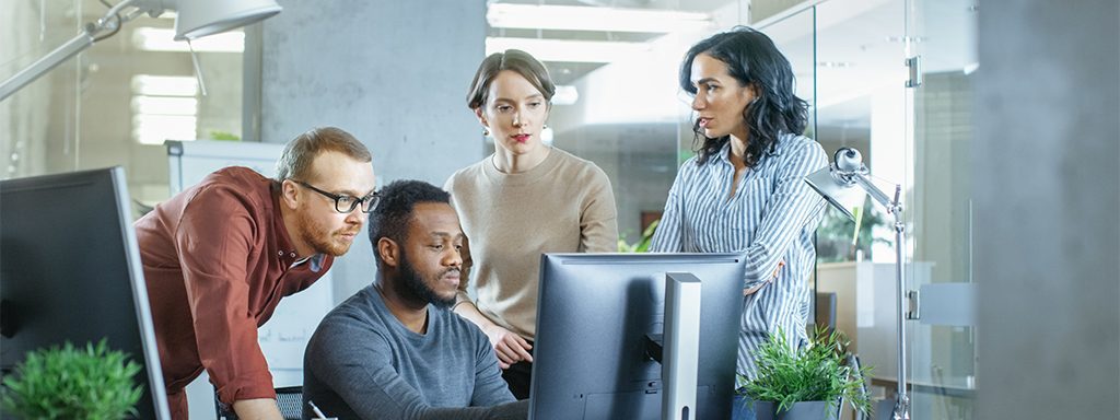 A man and women surrounding a man sitting next to the computer